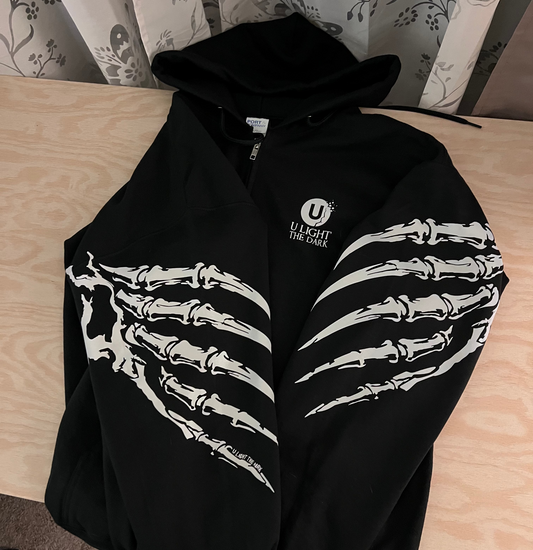A black zip-up hoodie laid flat on a light wood surface, featuring large white skeletal hand graphics across both forearms and a small white chest logo that reads 'U LIGHT THE DARK.' 